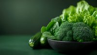 Fresh broccoli florets in black bowl with lettuce leaves and zucchini on dark green background, healthy eating concept with copy space.
