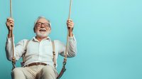 Happy senior Caucasian man with glasses and suspenders laughing while swinging on rope swing against turquoise background for retirement lifestyle concepts.