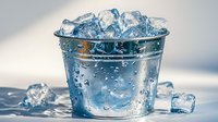 Clear glass bucket filled with ice cubes and water droplets against light background, fresh cold drink concept with condensation and reflective surface.