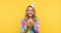 Happy young blonde woman in colorful tie-dye shirt and party hat holding birthday cupcake with candle, smiling at camera against bright yellow background.