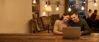 Young couple using laptop together in cozy cafe with warm lighting and vintage decor, sharing screen for work or entertainment in comfortable setting.