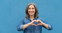 Happy mature Caucasian woman making heart shape with hands against blue wall background expressing love and positive emotions for lifestyle content.