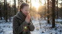 Young Caucasian woman in warm winter coat praying peacefully in snowy forest during golden hour sunrise with soft snowflakes falling around her.