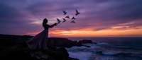 Woman in flowing dress releasing birds at dramatic ocean sunset with purple and orange sky creating magical silhouette scene for freedom and hope concepts.