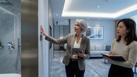 Professional mature Caucasian businesswoman presenting modern office space to young Asian female colleague during corporate real estate tour or workplace consultation.