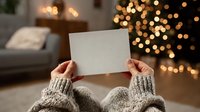 Hands in knitted sweater holding blank white card against blurred Christmas tree lights background in cozy living room setting.