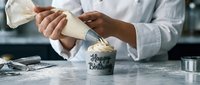 Pastry chef decorating a Happy Birthday cupcake with cream using a piping bag on a marble countertop in a professional bakery kitchen.