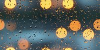Glowing bokeh lights through wet window glass with raindrops creating atmospheric mood. Night city view with golden circles and water droplets on dark background.