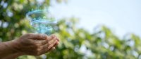 Hands holding clear glass of fresh water outdoors with green nature background. Hydration and healthy lifestyle concept for wellness campaigns.