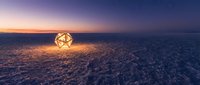 Glowing geometric lantern sits on vast desert landscape at twilight, casting warm light across sandy terrain during golden hour sunset.