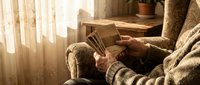 Senior man in cozy sweater reading book in comfortable armchair by window with warm sunlight streaming through curtains in peaceful home interior.