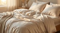 Unmade bed with beige silk or satin sheets and pillows in warm sunlight from window, showing wrinkled luxury bedding for home decor and lifestyle content.