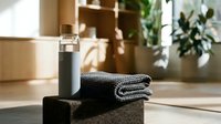 Sleek black cosmetic bottle with metallic cap displayed on stone platform alongside folded gray towels in modern spa or bathroom setting with wooden shelving and plants.