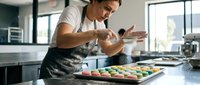Professional male chef in apron carefully decorating colorful macarons with precision in modern commercial kitchen for bakery production and culinary arts.