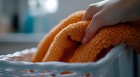 Orange microfiber towel being placed in laundry basket, close-up view showing soft texture and fabric detail in natural light, focus on hand movement.