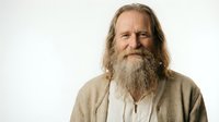 Mature Caucasian man with long gray beard and hair smiling warmly in casual linen shirt against white background for lifestyle and portrait photography.