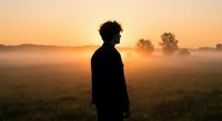 Young man silhouette standing in misty field during golden sunrise, peaceful morning landscape with fog and trees creating serene atmospheric mood.