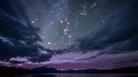 Dramatic night sky with Milky Way galaxy and stars over mountain silhouettes with purple clouds creating mystical atmosphere for astronomy and nature themes.