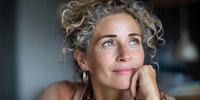 Mature woman with natural gray curly hair smiling thoughtfully with hand on chin, illuminated by soft natural light in contemplative moment.