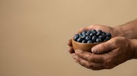 Senior man with dark skin holding wooden bowl filled with fresh blueberries against neutral background. Suitable for health, nutrition, organic produce, and wellness content.
