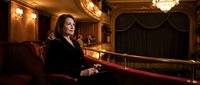 Elegant middle-aged Caucasian businesswoman in black suit sitting alone in luxurious vintage theater balcony with ornate golden architecture and red velvet seats.