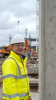 Senior construction manager in high visibility safety gear and hard hat standing at concrete building site with heavy machinery in background.