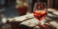 Glass of rosé wine on rustic wooden table with warm sunlight creating elegant shadows and bokeh background for wine tasting and dining concepts.