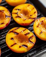Fresh halved peaches with caramelized grill marks on black iron grate, showing juicy flesh and charred surface texture in macro view. Perfect for summer recipes.