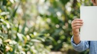 Young woman with light skin tone holding blank white card outdoors in natural garden setting, suitable for advertising, marketing campaigns, promotional materials, and business communications.