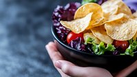 Fresh corn tortilla chips served with mixed lettuce, cherry tomatoes and purple cabbage in black ceramic bowl on dark background. Food photography for menu.