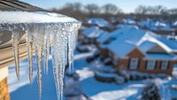 Dramatic icicles hanging from roof edge against blurred winter neighborhood background with snow-covered houses and bright blue sky. Seasonal weather concept.