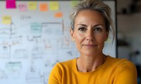 Confident mature businesswoman in yellow sweater smiling at camera, with sticky notes and planning board in background showing workplace organization and leadership.
