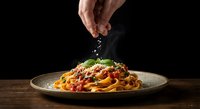 Hand sprinkling salt on delicious spaghetti pasta with tomato sauce and fresh basil leaves served on rustic plate against dark background.