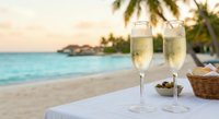 Two champagne flutes filled with sparkling wine on tropical beach table at sunset with palm trees and turquoise ocean creating romantic vacation atmosphere.