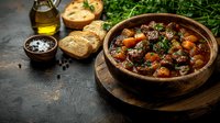 Rustic beef stew with carrots and herbs served in a wooden bowl with crusty bread, olive oil, and seasoning on dark slate surface for cooking and food styling.