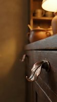 Rustic wooden cabinet with ornate metal leaf handle in warm pottery studio workshop interior with ceramic vessels and soft lighting atmosphere.