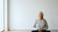 Senior woman with long white hair and light complexion, wearing a gray knit sweater, sitting cross-legged on floor against white wall. Meditation and wellness concept.