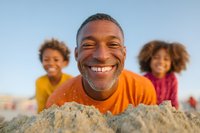 African American father with bright smile lying on sandy beach with two playful children in colorful t-shirts enjoying summer family vacation outdoors on sunny day.