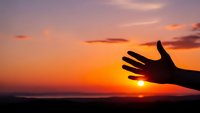 Human hand silhouette reaching toward golden sunset sky with vibrant orange and pink clouds over horizon for inspirational and spiritual concepts.