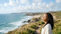 Young woman with dark braided hair and medium skin tone standing on coastal cliff overlooking rocky shoreline and ocean waves, wearing white shirt, suitable for travel, lifestyle, and wellness content.