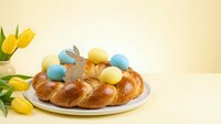 Traditional Easter braided bread with colorful eggs and wooden bunny decoration alongside yellow tulips on neutral background for holiday celebrations.