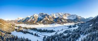 Aerial view of snow-covered alpine valley with forested slopes and dramatic mountain peaks under clear blue sky, suitable for travel and nature backgrounds.