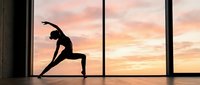 Young woman practicing yoga side stretch pose silhouette against large windows with pink sunset sky background for wellness and fitness lifestyle.