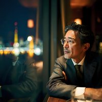 Mature Asian businessman in suit contemplating city skyline through office window at night with urban lights creating atmospheric corporate setting.