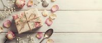Vintage gift box wrapped in kraft paper with twine bow surrounded by dried rose petals and baby breath flowers on rustic white wooden background.
