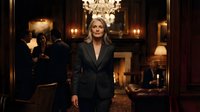 Professional blonde businesswoman in elegant black suit standing confidently in luxury hotel lobby with crystal chandelier and warm ambient lighting.