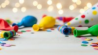 Colorful party supplies with balloons, streamers and confetti scattered on white surface with warm bokeh lights creating festive celebration atmosphere.