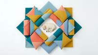 Newborn baby sleeping peacefully in center of colorful geometric fabric arrangement with teal, coral, yellow and blue diamond-shaped cloth pieces overlapping on white background.