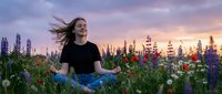 Young woman with flowing hair meditating peacefully in wildflower meadow at golden sunset, surrounded by colorful lupines and blooms in serene natural setting.