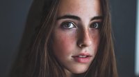 Young brunette woman with freckles and natural makeup looking directly at camera with soft lighting for beauty and lifestyle content.